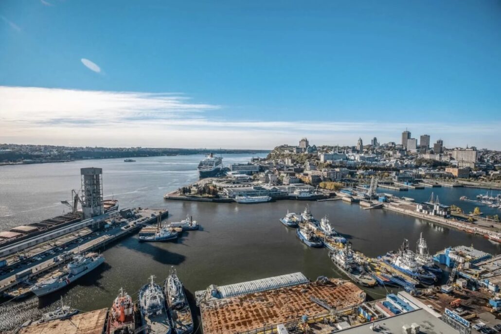 Aerial view of the Port of Quebec with multiple docked ships, including the Brilliant Lady on her Maiden Call. Industrial buildings and a city skyline rise in the background under a clear blue sky, with urban structures surrounding the waterfront.