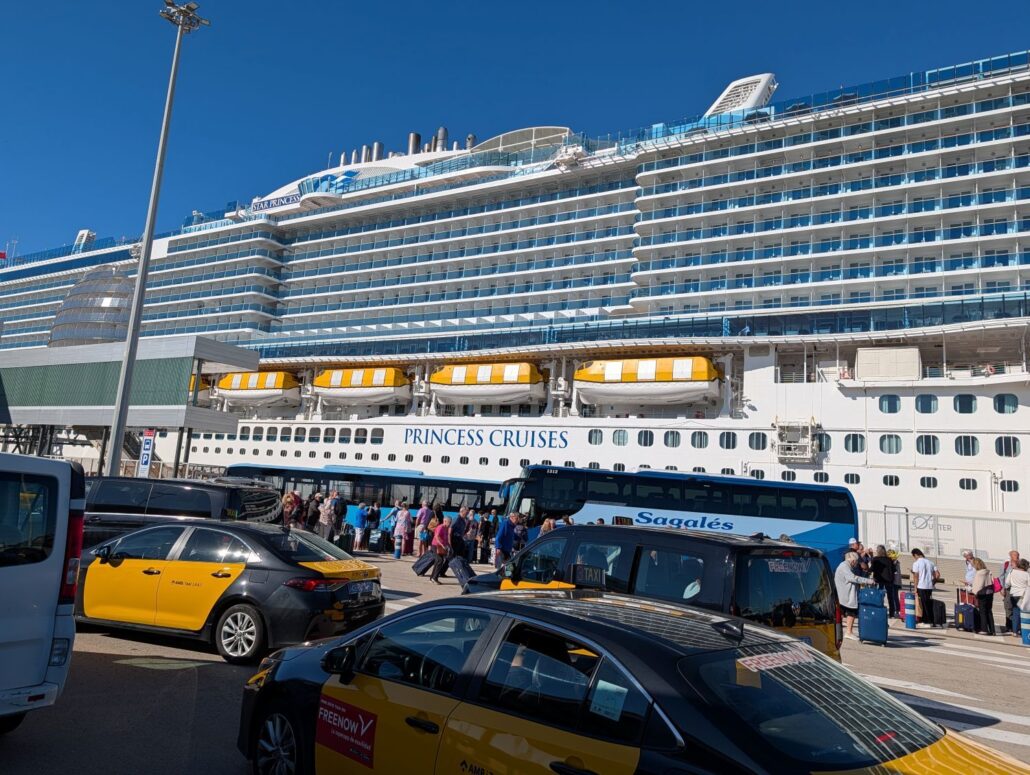 A large cruise ship labeled "Princess Cruises," the Star Princess, is docked at a port. People are lined up on the pavement near buses and taxis under clear blue skies. The ship’s lifeboats and multiple decks are visible.