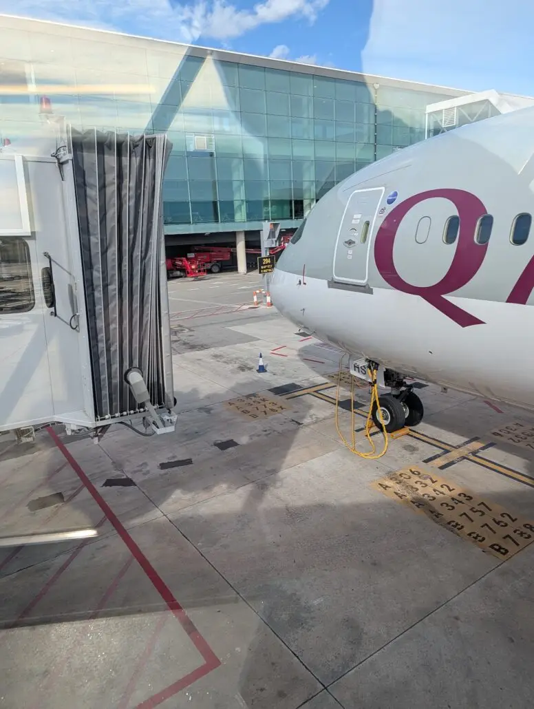 A Qatar Airways airplane is parked at an airport gate, connected to a jet bridge. The ground is marked with parking spots, while a glass terminal building, reminiscent of those seen on an Inaugural Cruise with Star Princess, stands in the background.