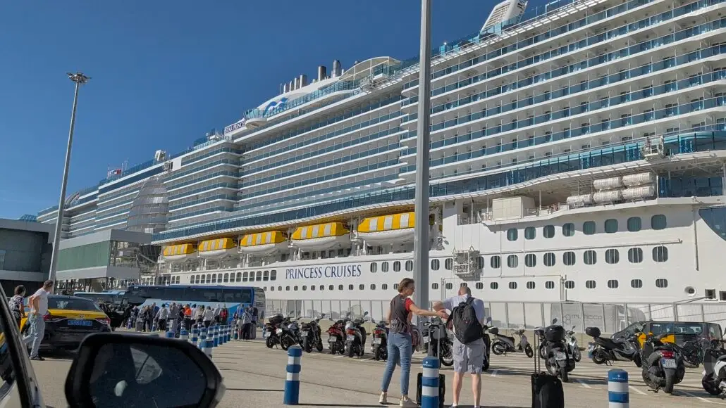 A large cruise ship labeled "Star Princess" is docked at a port. People stand near parked motorcycles, cars, and buses under a clear blue sky, with a tall lamppost prominent in the foreground.