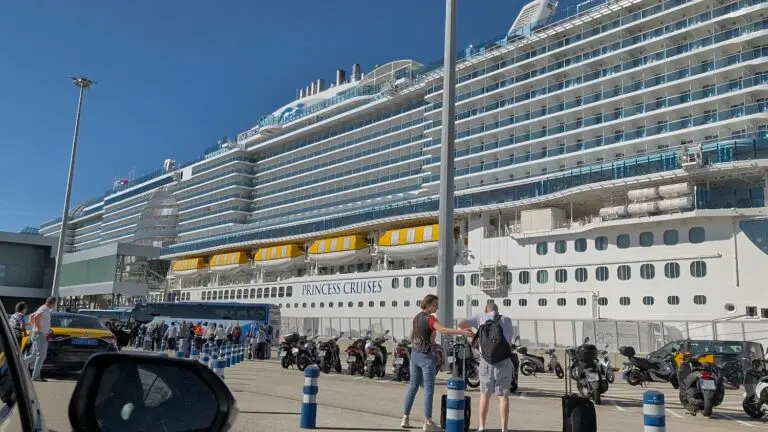 A large cruise ship labeled "Star Princess" is docked at a port. People stand near parked motorcycles, cars, and buses under a clear blue sky, with a tall lamppost prominent in the foreground.