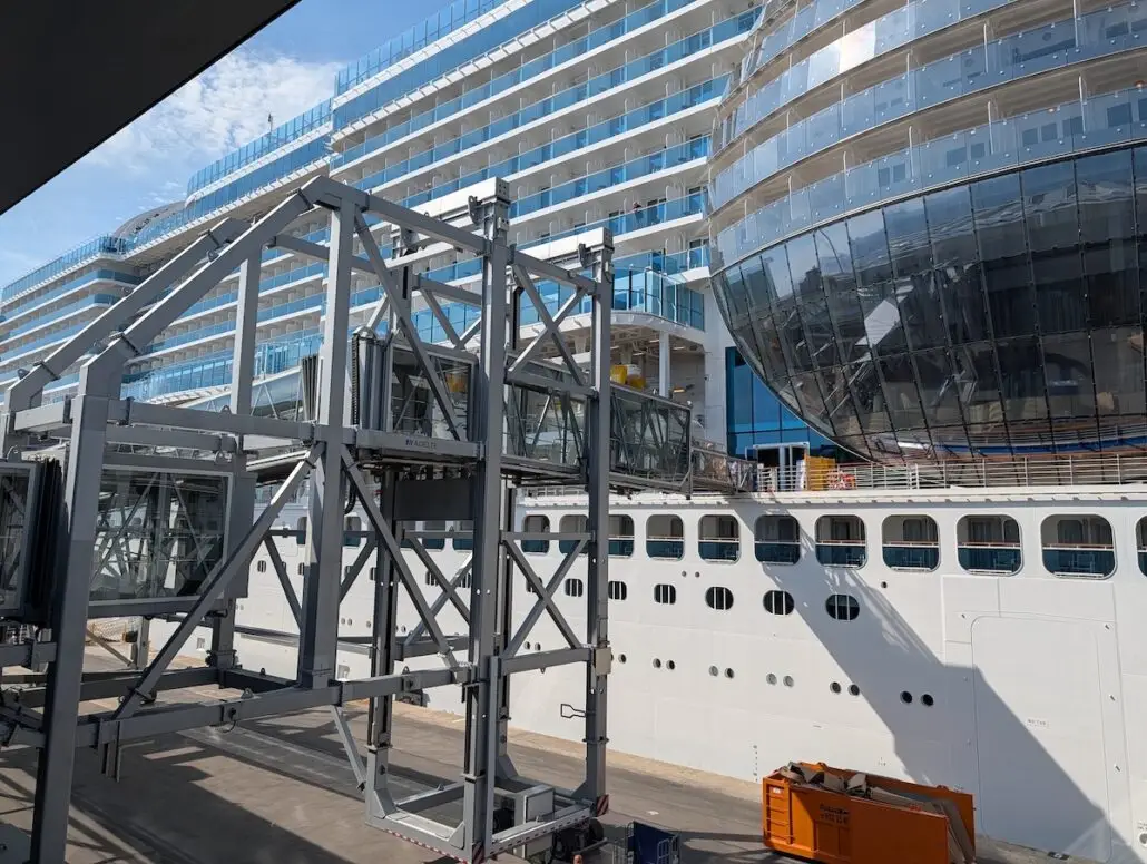 A large white cruise ship, the Star Princess, is docked at the port of Barcelona, with glass boarding bridges connected to its side. Part of a modern spherical structure and some equipment are visible in the foreground.