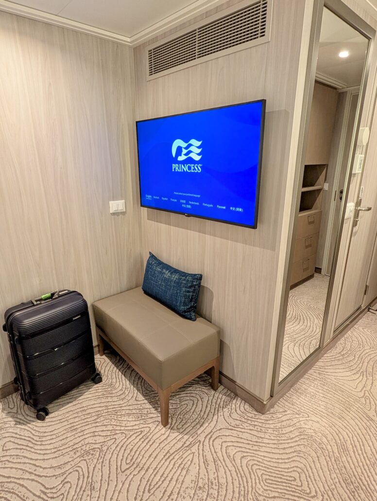 A small Star Princess cruise ship cabin area with a suitcase on the floor, a padded bench with a blue pillow, and a wall-mounted TV displaying the Princess Cruises logo. A mirror and shelves are visible, ready for your Transatlantic Inaugural Cruise boarding.