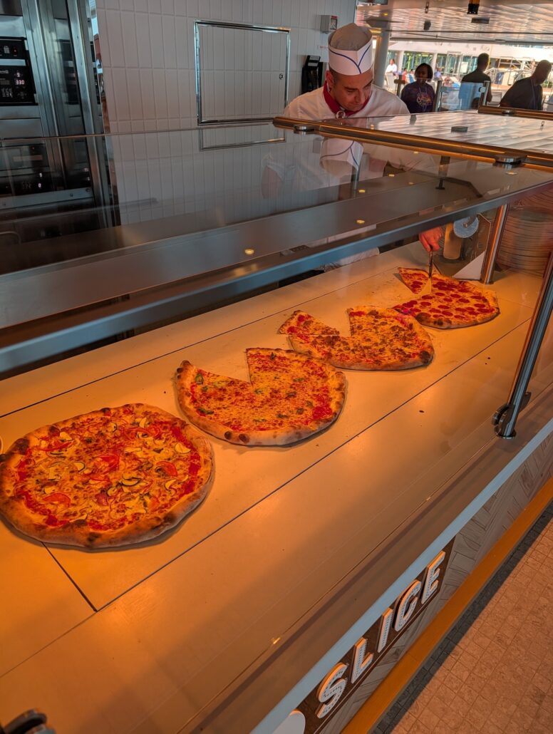 A worker in a white uniform and cap stands behind a glass counter displaying four large pizzas, one with several slices missing. The spotless counter and kitchen set the scene for dining on the Star Princess during its inaugural cruise.