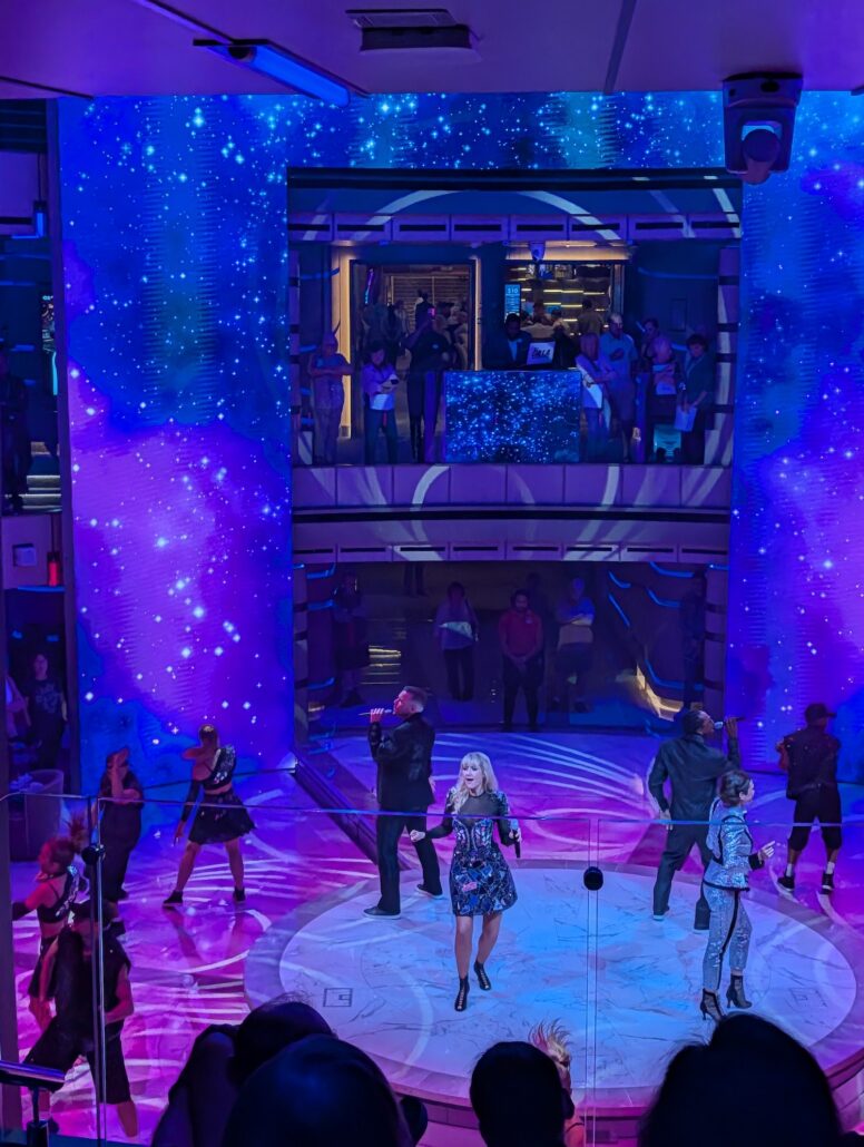 A group of performers sing and dance on a circular stage with blue and purple galaxy-themed lighting aboard the Star Princess, while guests watch from balconies above in a multi-level indoor venue during the inaugural cruise.
