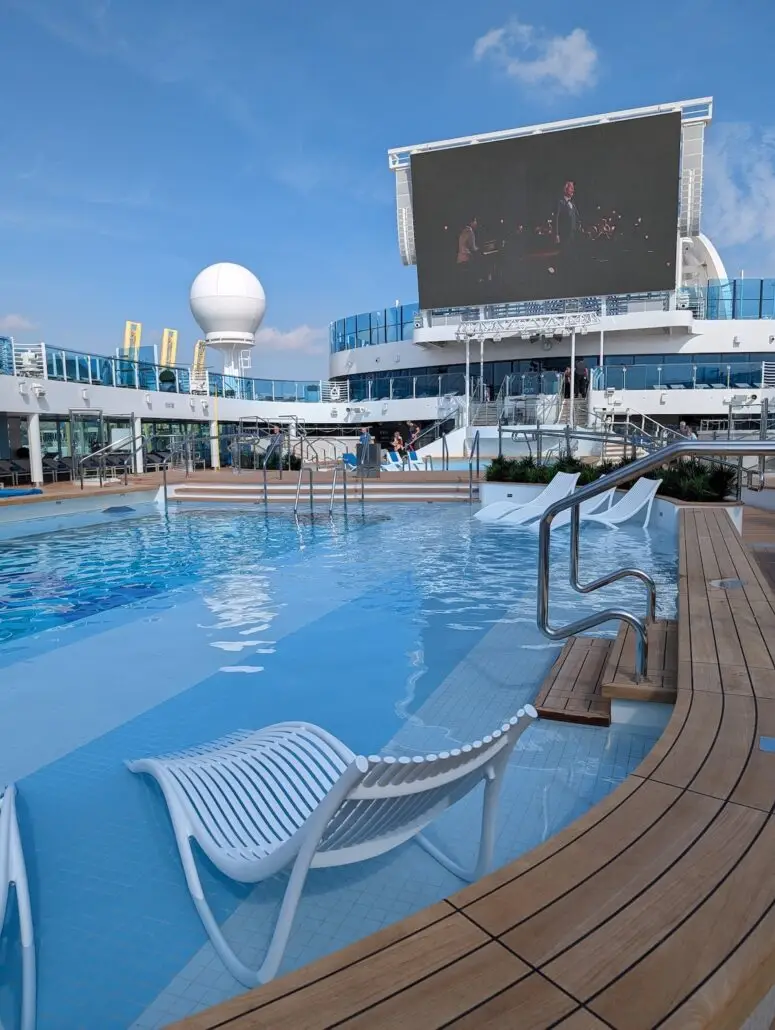 A swimming pool on the deck of the Star Princess during its maiden voyage, featuring white lounge chairs partially in the water, metal railings, and a large outdoor screen. Blue sky and ship structures are visible as Barcelona fades into the background.