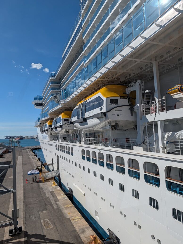 The Star Princess, a large white cruise ship, is docked at the port for its Inaugural Cruise, displaying multiple decks with balconies, yellow lifeboats along the side, a blue sky, and part of the port infrastructure.