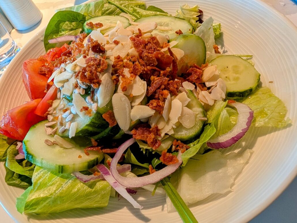 A fresh salad with sliced cucumbers, tomatoes, red onions, leafy greens, sliced almonds, sunflower seeds, and crumbled bacon graces a white plate—perfect for a light meal aboard the Star Princess during your Transatlantic Cruise. A glass of water sits nearby.
