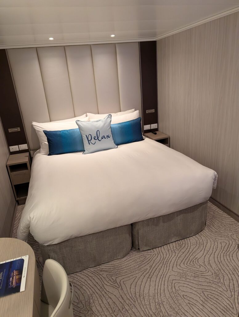 A modern hotel room with a neatly made double bed, white bedding, blue accent pillows, and a decorative “Relax” pillow—perfect for unwinding before or after your Star Princess Transatlantic cruise. Nightstands, lamps, and a cozy chair complete the space.