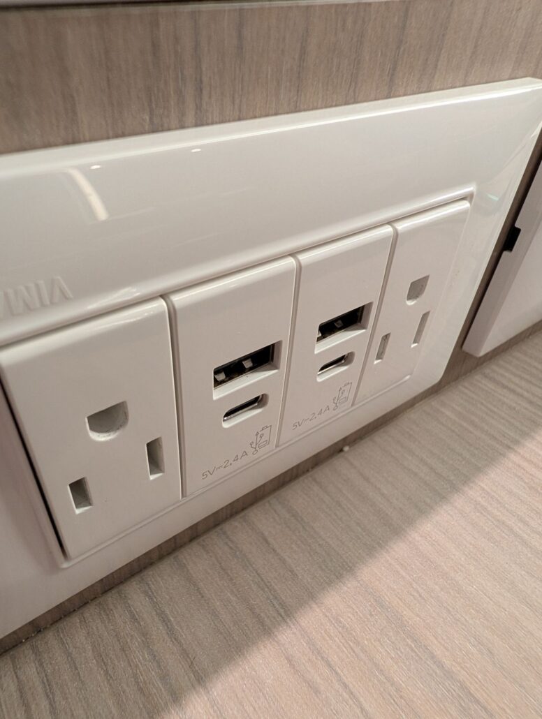 A close-up view of a wall outlet panel with two standard electrical sockets and two USB charging ports, installed on a light wood wall surface aboard the Star Princess during its Transatlantic Inaugural Cruise.