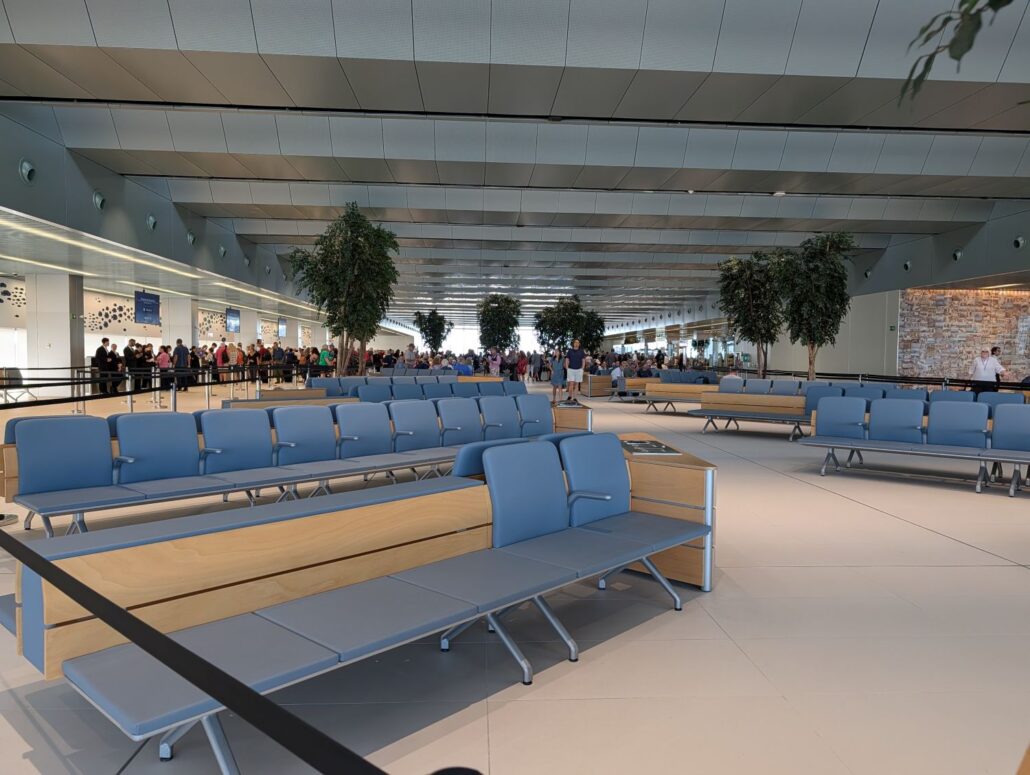 Rows of empty blue and wooden seats in a modern airport waiting area evoke the anticipation of a Star Princess inaugural cruise, with potted trees and a high ceiling. In the background, groups of people stand in line near counters.