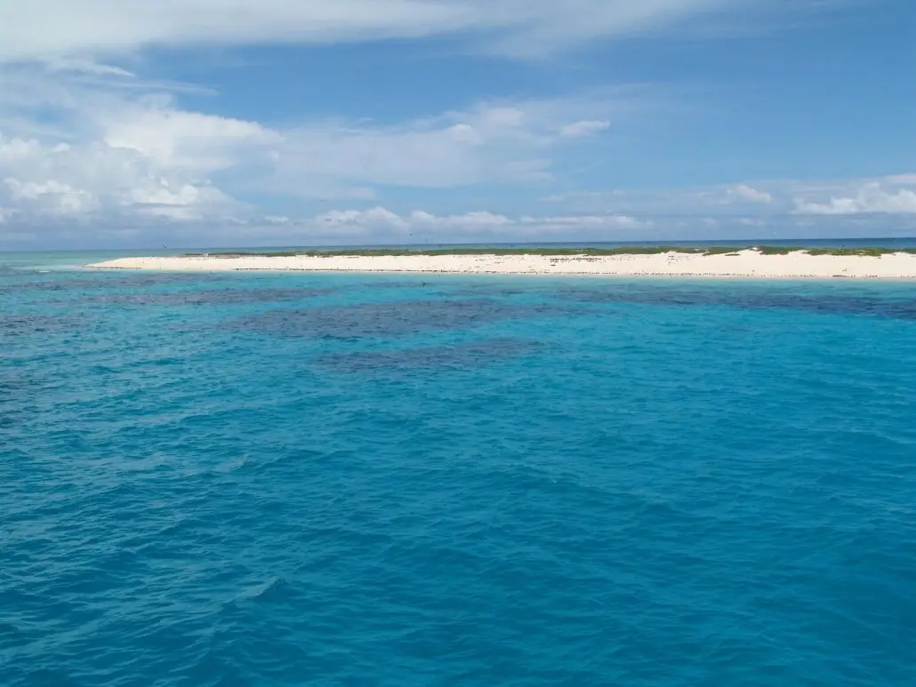 Clear turquoise ocean water in the foreground with a white sandy beach and low green vegetation on a narrow strip of land in the background under a partly cloudy blue sky, where a cruise guest was left behind on the island.