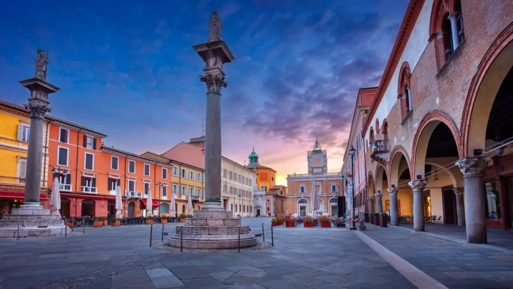A town square at sunset with colorful buildings, two stone columns with statues, arched walkways, and a clock tower in the background under a partly cloudy sky, reminiscent of an Italian cruise terminal welcoming American travelers.