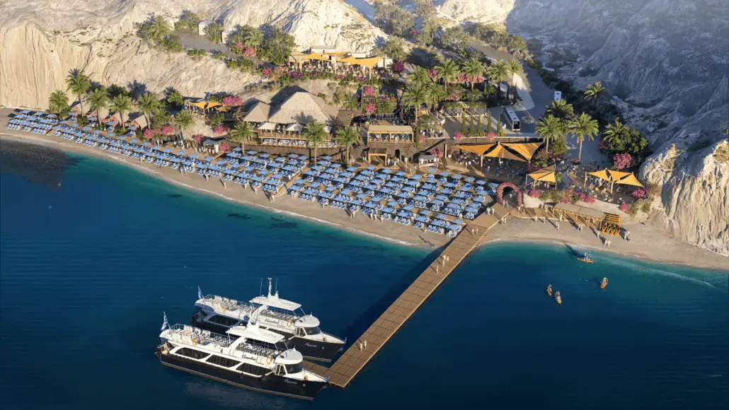 Aerial view of Royal Beach Club Santorini, opening Summer 2026, with rows of blue lounge chairs and umbrellas, palm trees, multiple buildings, a long wooden dock, two yachts moored for cruise guests, and kayaks near rocky cliffs.