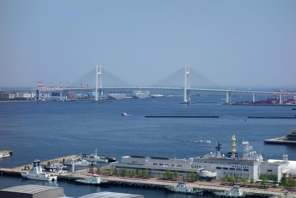 A wide river with several boats, bordered by docks and buildings—some serving new Asian ports—features a large cable-stayed bridge spanning the water in the background under a clear sky.