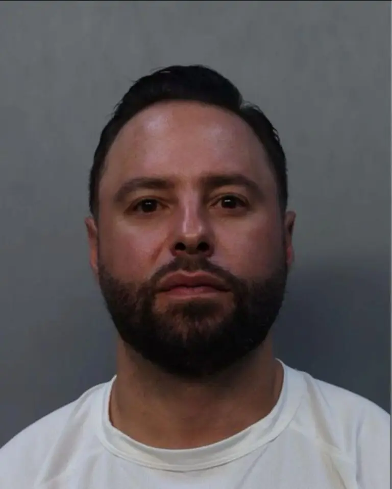A man with short dark hair and a beard is facing forward against a plain gray background, wearing a white shirt—appearing much like a defendant in cruise ship assault charges handled by Florida lawyers.