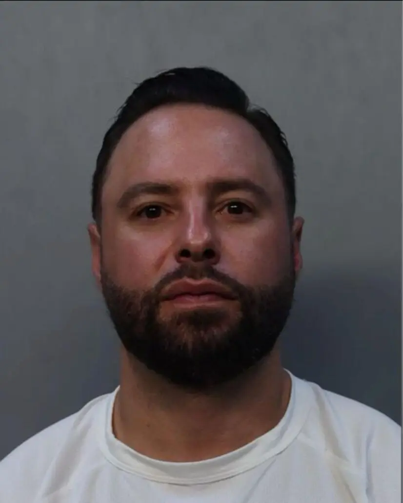 A man with short dark hair and a beard is facing forward against a plain gray background, wearing a white shirt—appearing much like a defendant in cruise ship assault charges handled by Florida lawyers.