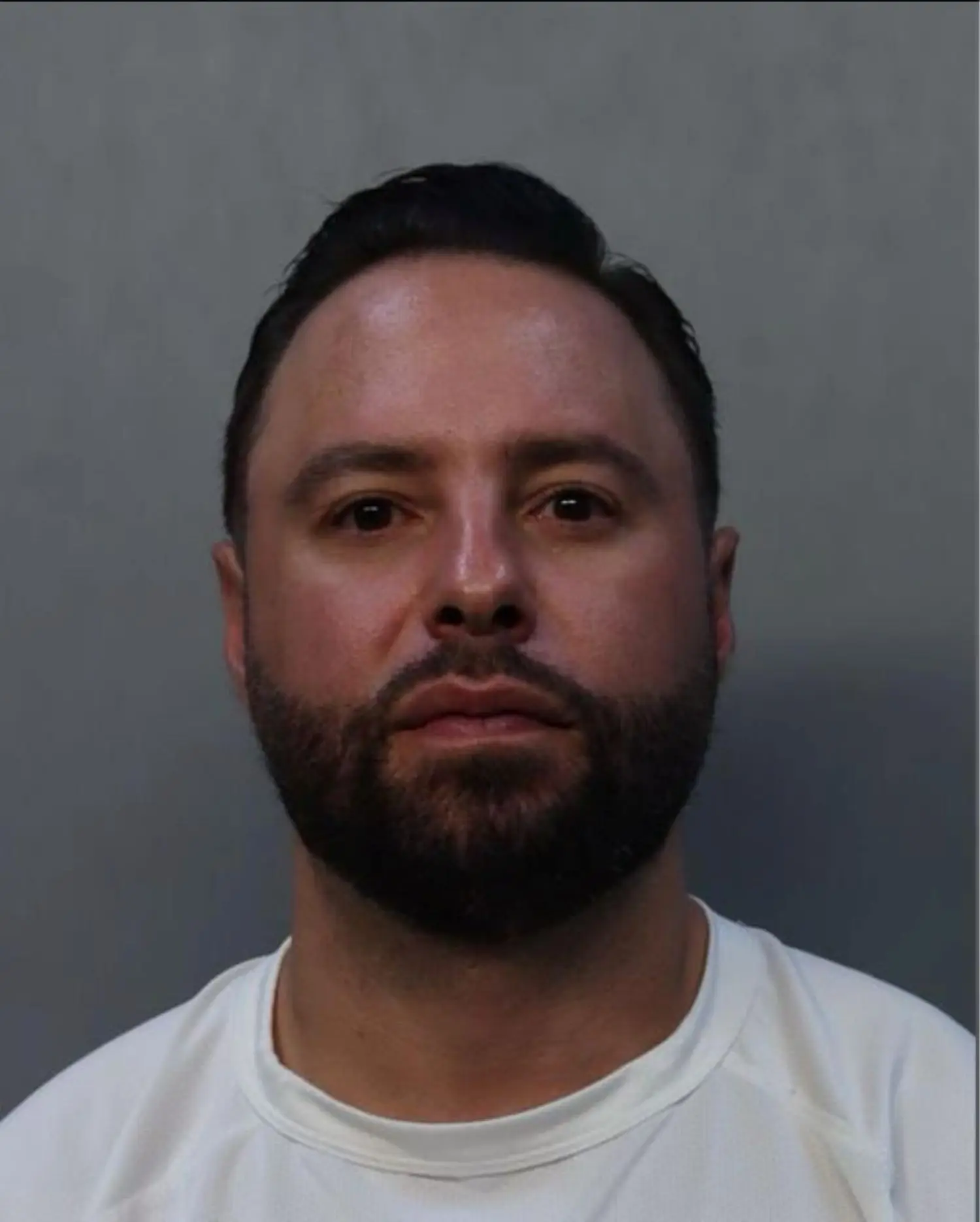 A man with short dark hair and a beard is facing forward against a plain gray background, wearing a white shirt—appearing much like a defendant in cruise ship assault charges handled by Florida lawyers.