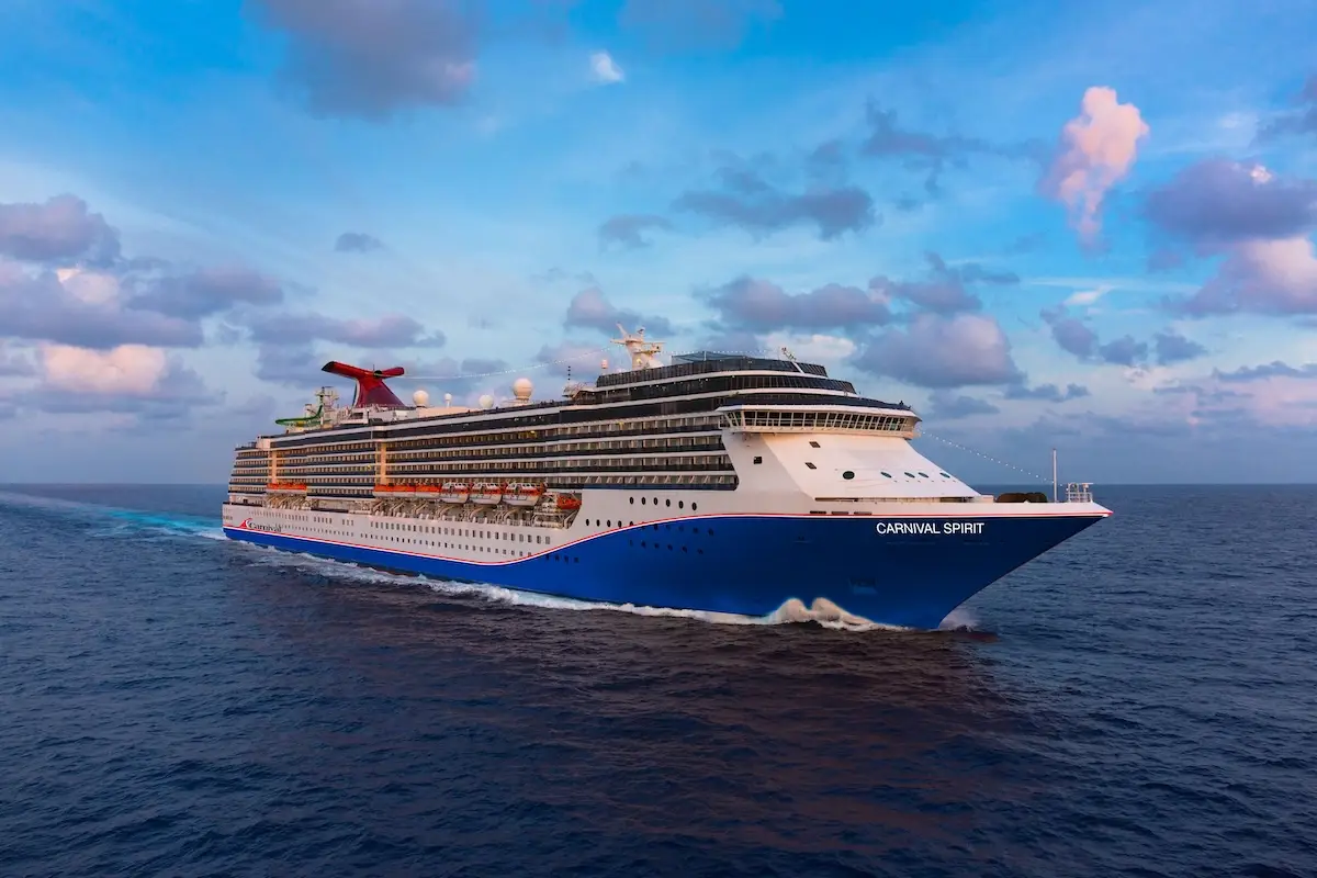 A large Carnival cruise ship named Carnival Spirit sails across calm ocean waters under a partly cloudy sky during daytime, showcasing multiple decks, lifeboats, and its distinctive red funnel. Experience adventure on the Carnival Spirit Tampa voyage.