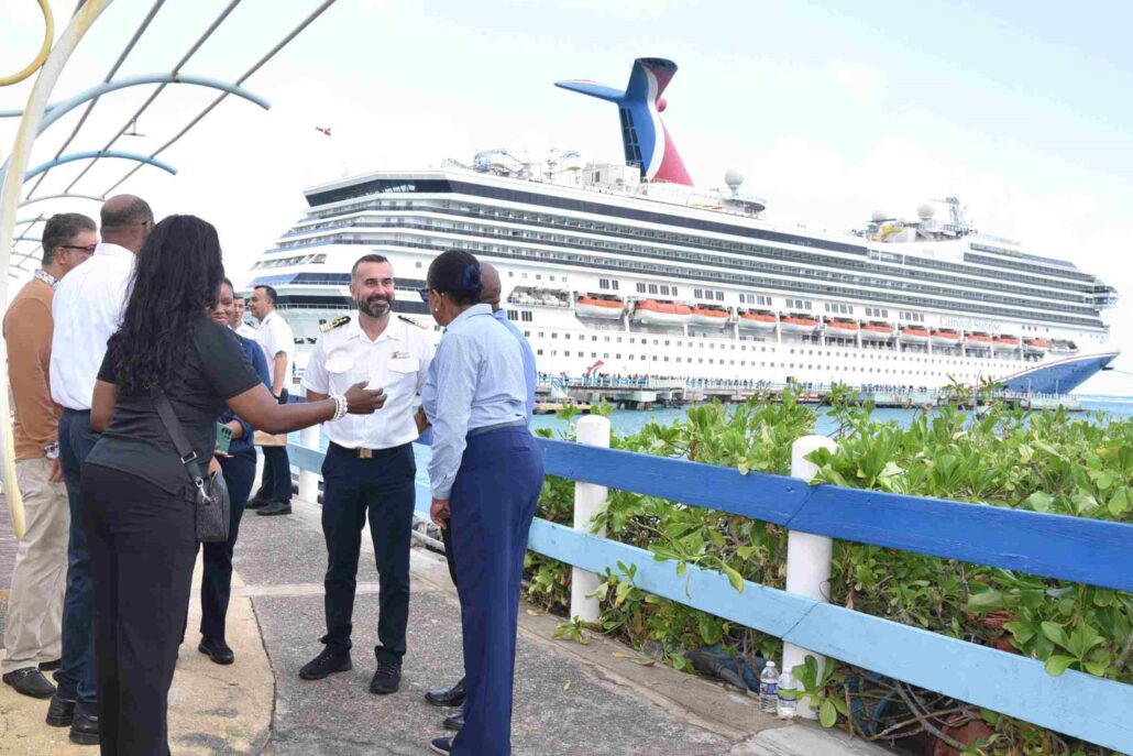 A group of people, including a man in a white uniform, stand near a railing having a conversation by a dock in Ocho Rios, with a large Carnival Cruise ship in the background under partly cloudy skies.