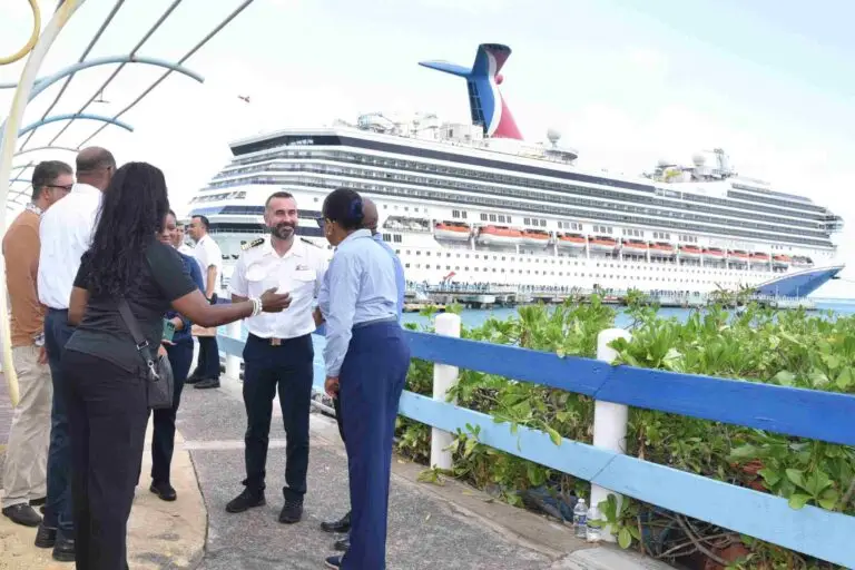 A group of people, including a man in a white uniform, stand near a railing having a conversation by a dock in Ocho Rios, with a large Carnival Cruise ship in the background under partly cloudy skies.