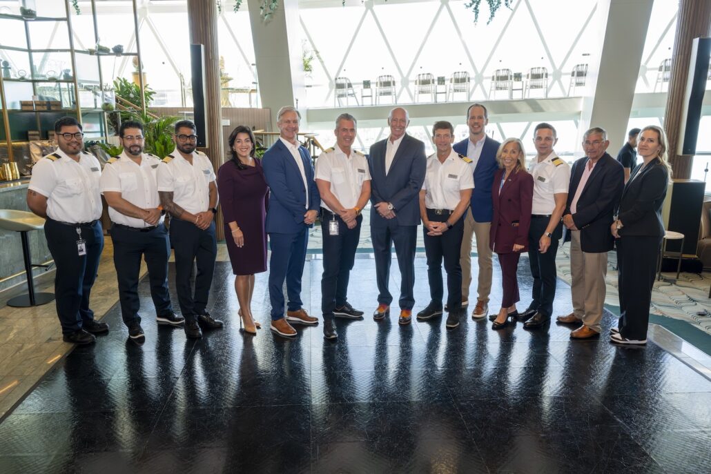 A group of thirteen people, including several in pilot uniforms and others in business attire, stand in a row inside a modern, well-lit building with large windows—capturing the excitement of Florida's Space Coast before embarking on Celebrity Apex Seven-Night Cruises.