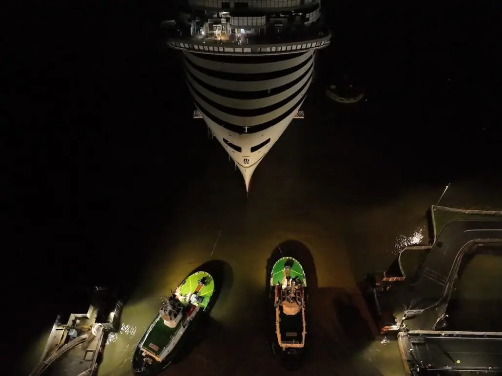 A large cruise ship docked at night, viewed from above, with its bow facing the camera. Two small tugboats with green decks are positioned near the front of the ship, illuminated by artificial lights.