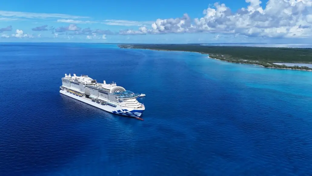 The Star Princess, a large white cruise ship, sails near a tropical coastline with clear blue water and lush green landscape under a bright sky, offering nightly entertainment before arriving at sun-soaked Princess Cay.