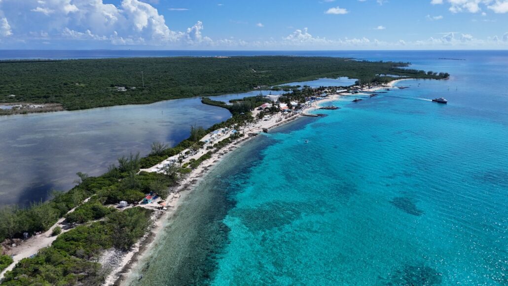 Aerial view of Princess Cay’s coastline with clear turquoise water, white sandy beaches, and lush vegetation. A few boats are near the shore, and scattered buildings line the beach under a blue sky as Star Princess visits this scenic paradise.