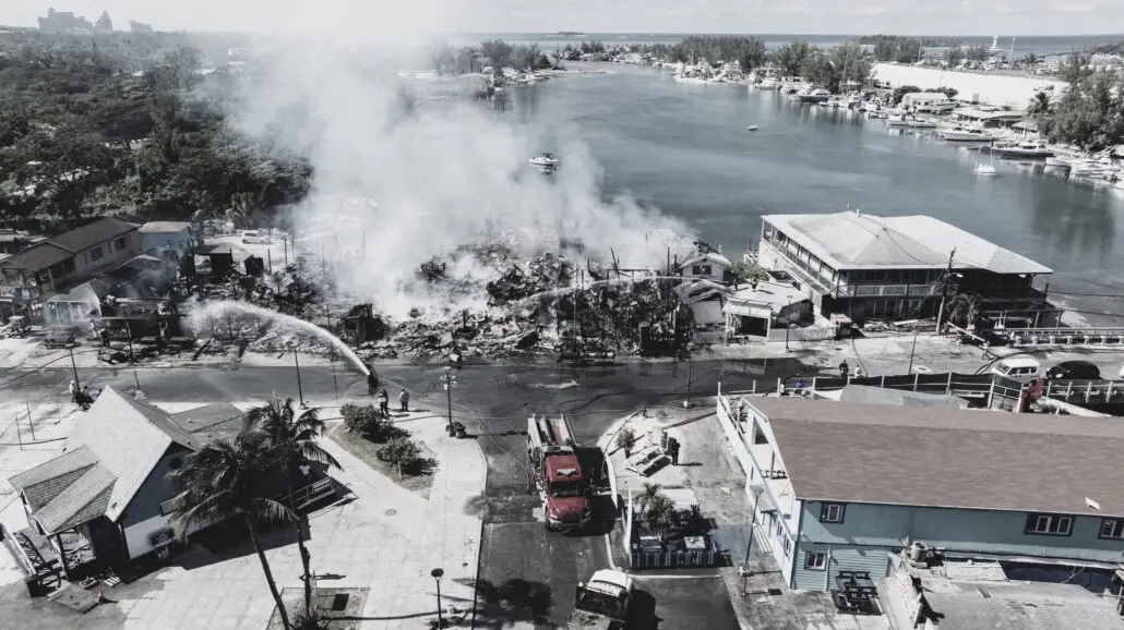 A large building fire near a waterfront is being extinguished by firefighters. Heavy smoke rises from the ruins, and fire trucks are parked on the street. Boats are visible in the water and nearby buildings appear undamaged.