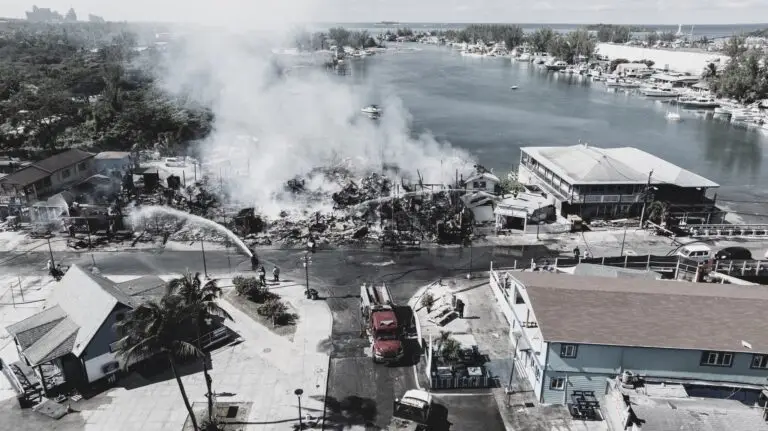 A large building fire near a waterfront is being extinguished by firefighters. Heavy smoke rises from the ruins, and fire trucks are parked on the street. Boats are visible in the water and nearby buildings appear undamaged.