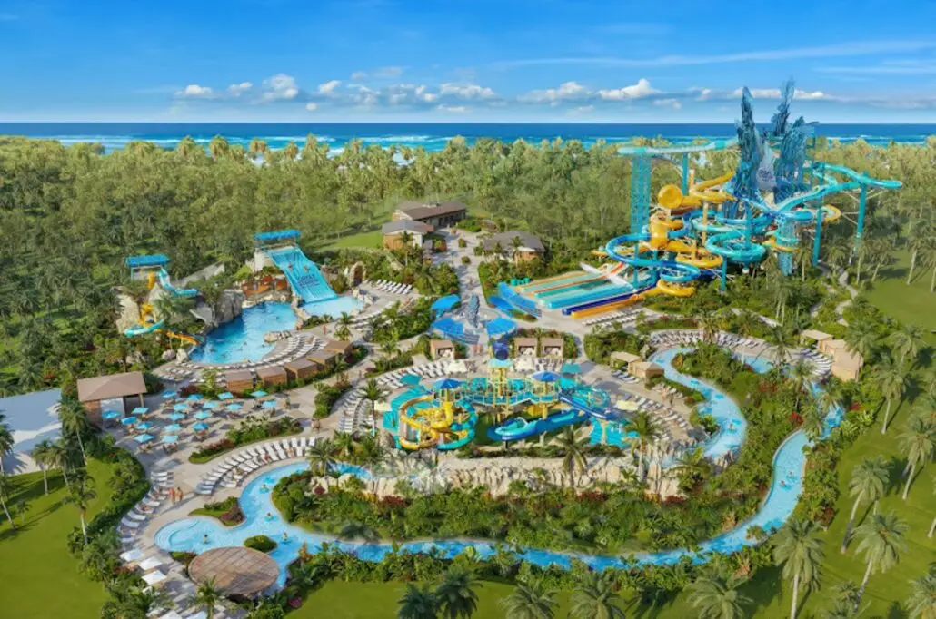 Aerial view of a large water park on the Norwegian Luna, featuring multiple pools, water slides, a lazy river, lounge chairs, tropical landscaping, and nearby forested areas—with the ocean visible in the background.