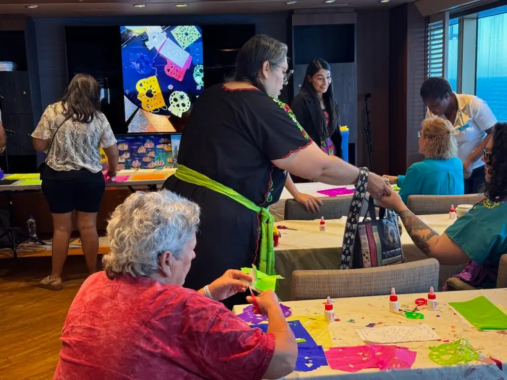 People seated and standing around tables participate in an arts and crafts activity with paper, glue, and scissors during one of Holland America’s Cultural Workshops. A large screen displays colorful decorative designs in a lively community room or classroom.