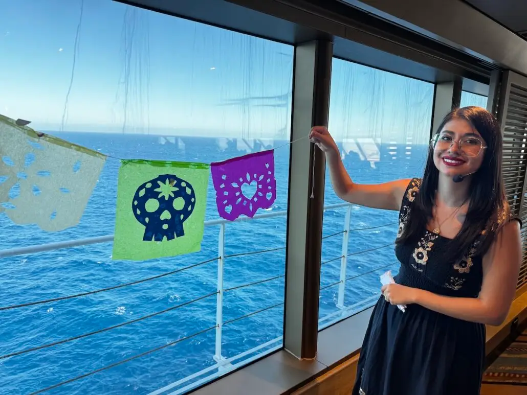 A woman in a black dress stands indoors by a large window overlooking the ocean, holding colorful papel picado banners with skull and heart designs—a vibrant scene from Holland America’s Mexico Cruises cultural workshops. Sunlight streams in through the window.