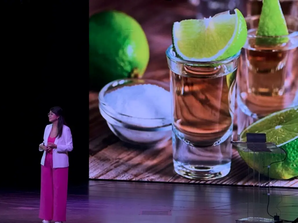 A woman in a pink outfit stands on stage during a Cultural Workshop on a Holland America Mexico Cruise, in front of a large screen displaying two tequila shots with lime wedges and a bowl of salt. A clear podium with a laptop is to her right.
