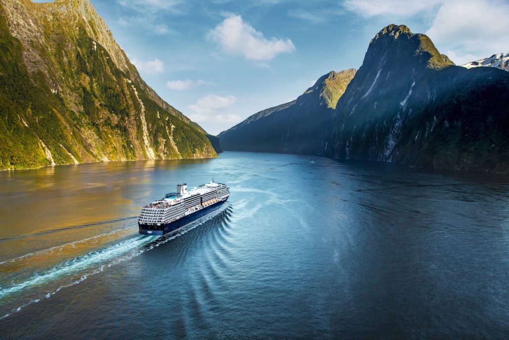 A large Holland America cruise ship sails through a wide, calm fjord surrounded by steep, green mountains under a partly cloudy sky. The water reflects the landscape and the ship, known for its overnight ports, leaves a gentle wake behind.