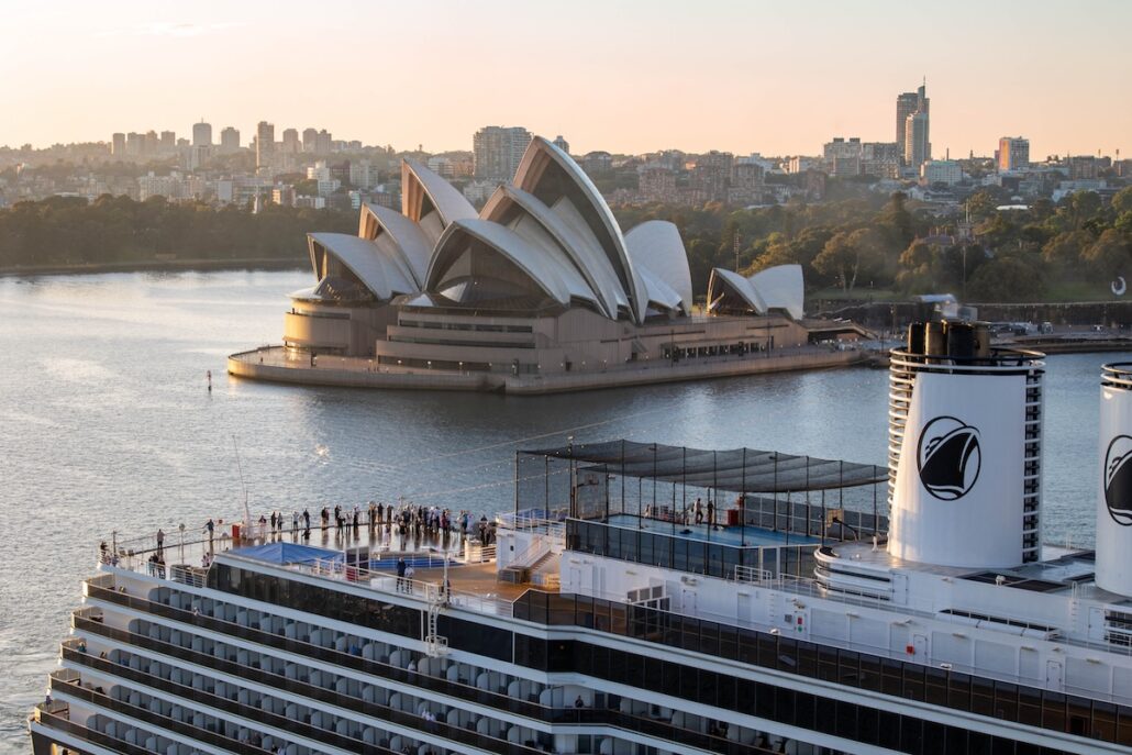 A large Holland America 2027-28 cruise ship is docked in front of the Sydney Opera House with a cityscape and calm water at sunset. Tourists are visible on the upper deck, enjoying one of the Overnight Ports Australia experiences.
