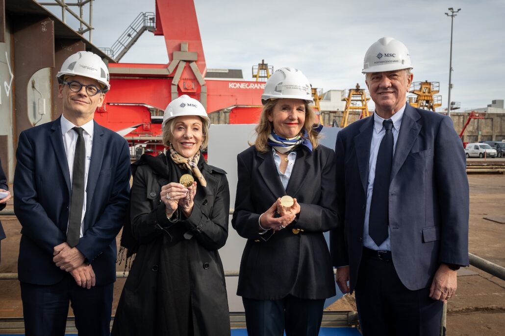 Four adults in business attire and white hard hats stand side by side at an industrial site. Two of them are holding circular objects. Industrial equipment and cranes are visible in the background.