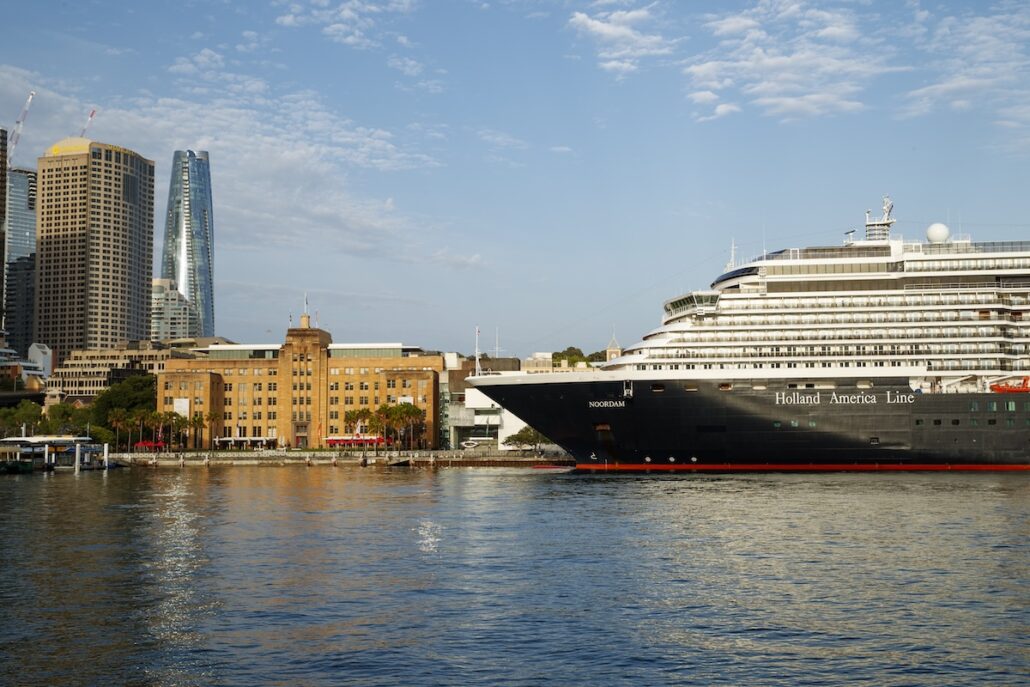 A large Holland America Line cruise ship is docked near a waterfront with modern high-rise buildings and an older brick building in the background under a partly cloudy sky.