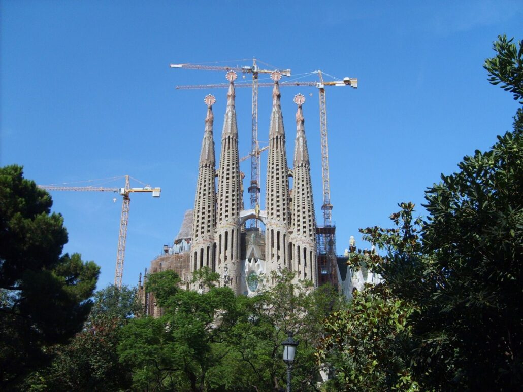 The Sagrada Família basilica in Barcelona, Spain, stands with multiple construction cranes around its spires—a must-see for first-time visitors and a highlight of Spain tourism. The clear blue sky and green trees beautifully frame the scene.