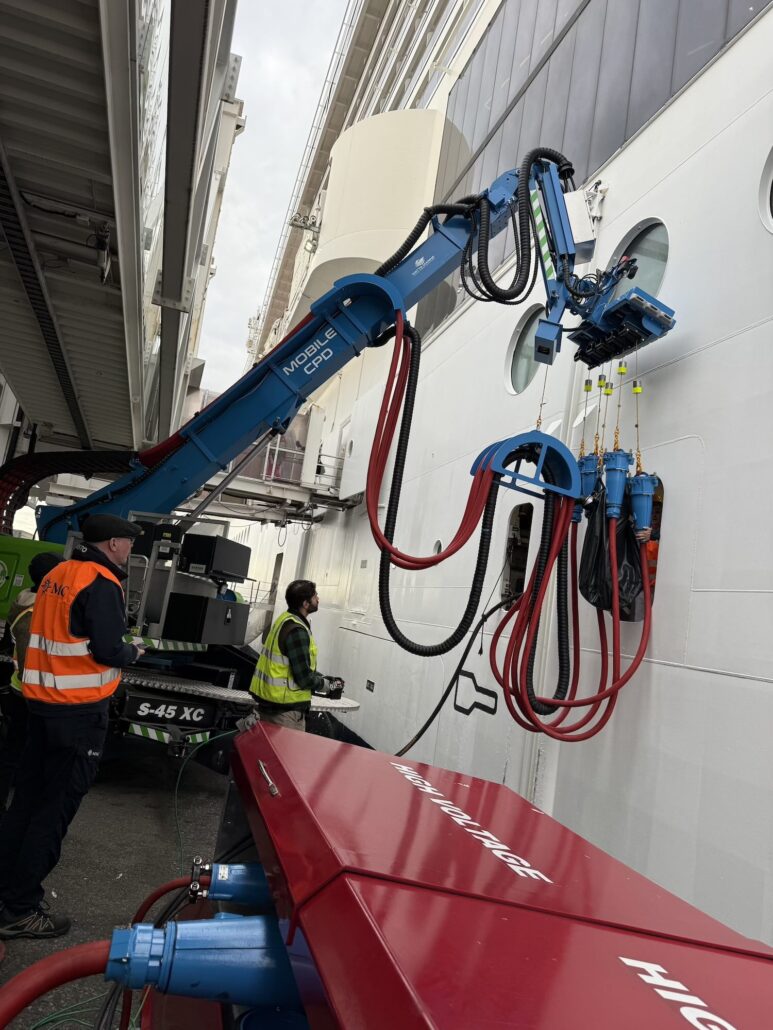 A blue robotic arm with suction pads is attached to the side of the MSC Meraviglia in NYC. Two workers in safety vests monitor the shore power machinery on the dock, while thick red and black hoses hang from the device.