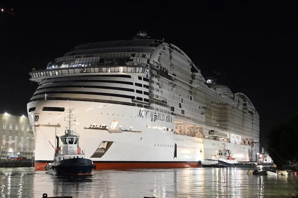 A large cruise ship, MSC World Asia, docked at night with lights on. A small tugboat is near the ship's bow, with water reflecting the lights and part of a building visible in the background.