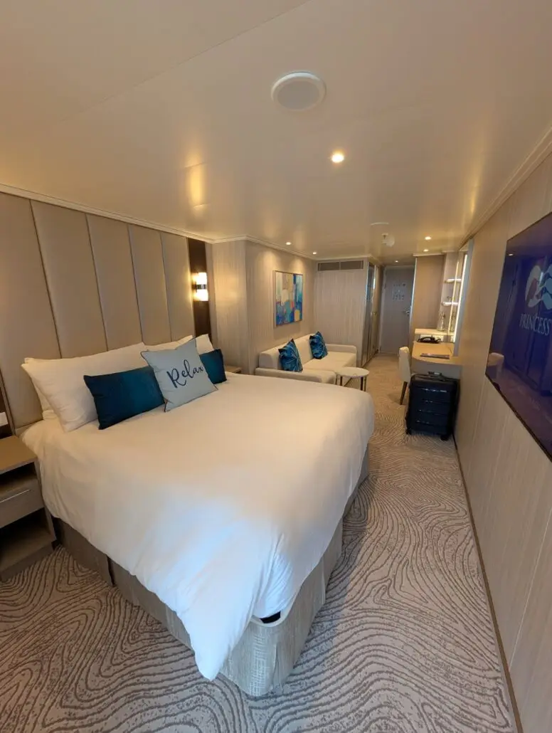 Modern cruise ship cabin with a large bed, blue accent pillows, small sofa, desk with mirror, wall art, and a TV displaying the Princess Cruises logo. Enjoy nightly entertainment and delicious food after exploring destinations like Princess Cay.