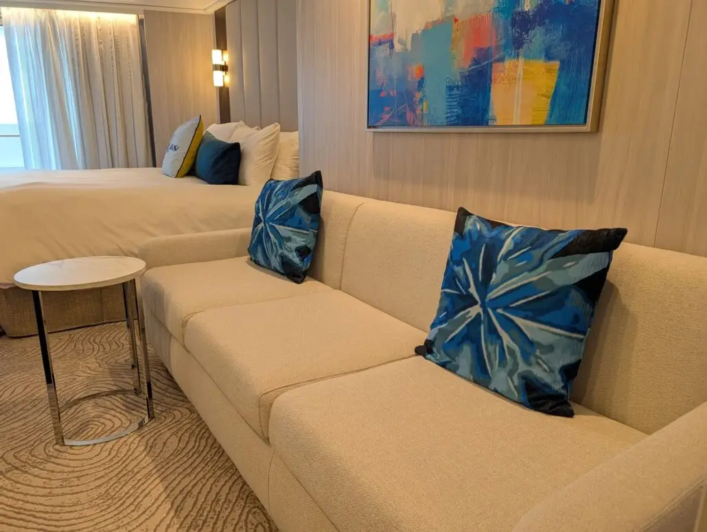 A beige sofa with two blue patterned throw pillows sits next to a small round side table, perfect for snacks or food. In the background, a bed with white bedding and blue pillows, a modern wall lamp, and an abstract painting complete the scene.