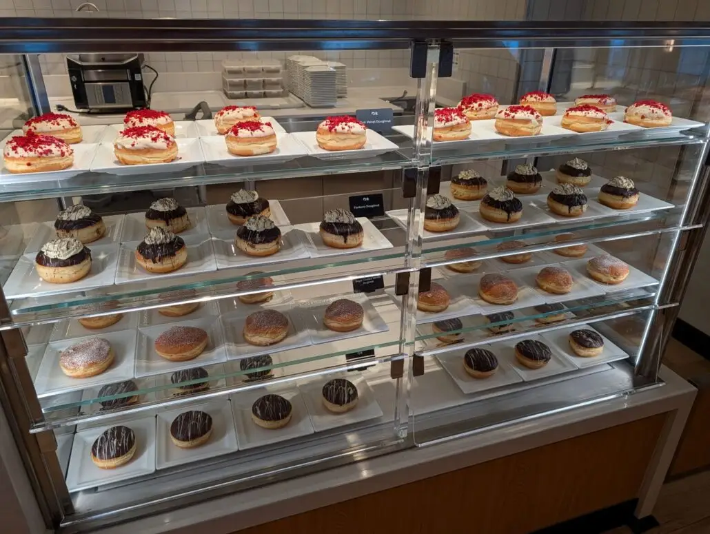 A glass display case with four shelves, each holding rows of neatly arranged donuts and pastries on white plates. Perfect for a sweet treat at Princess Cay, these desserts are a highlight among the food offerings during nightly entertainment.