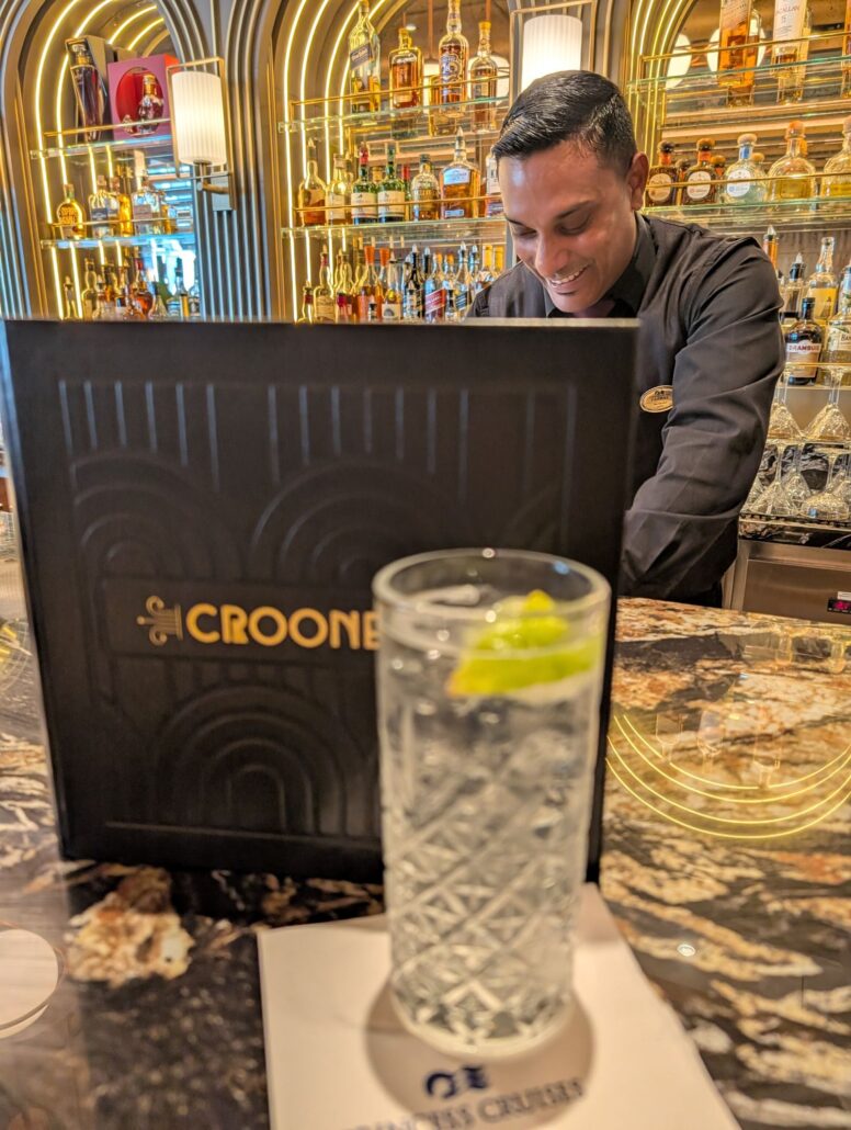 A bartender stands behind a bar with shelves of liquor bottles. In the foreground, a cocktail garnished with lime sits on a napkin next to a menu labeled “Crooner’s,” inviting guests to savor food and enjoy nightly entertainment.
