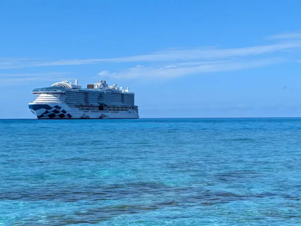 A large white cruise ship is anchored on a calm, blue ocean near Princess Cay under a mostly clear sky, with gentle waves and some shallow, turquoise water visible in the foreground.