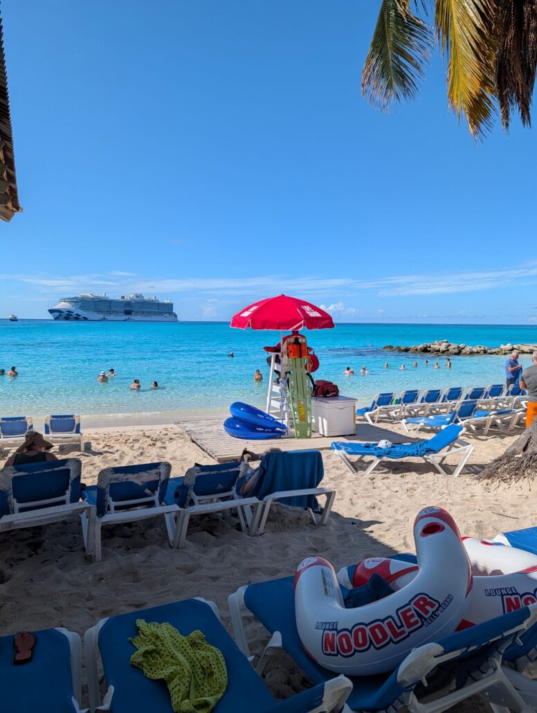 A beach scene at Princess Cay with rows of blue lounge chairs, a lifeguard stand under a red umbrella, inflatable pool floats, people swimming in clear blue water, and a cruise ship in the distance. Palm leaves hang overhead.
