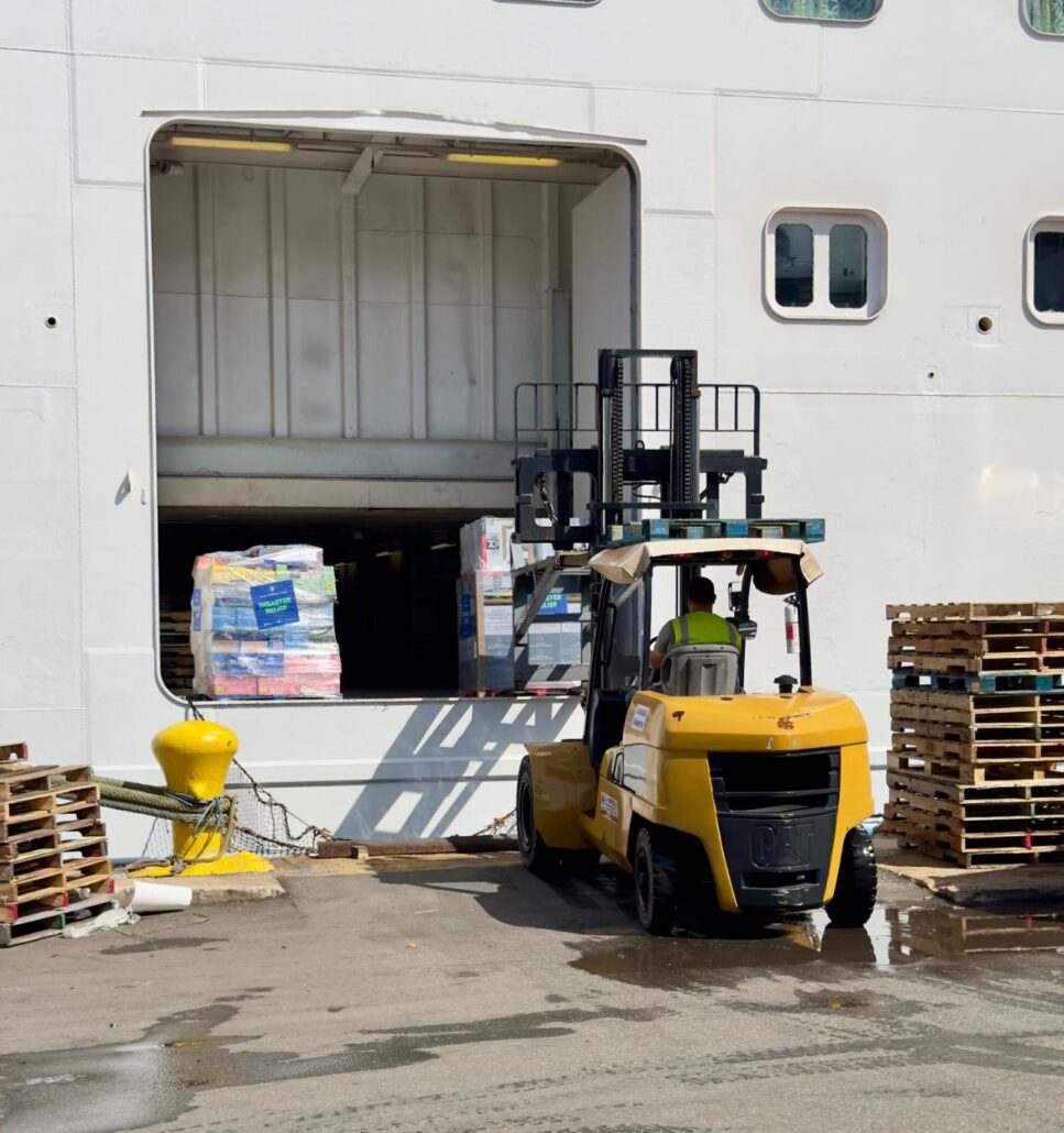 A yellow forklift moves pallets loaded with boxes into a rectangular opening on the side of a large white ship. Stacks of empty wooden pallets are nearby on the dock.