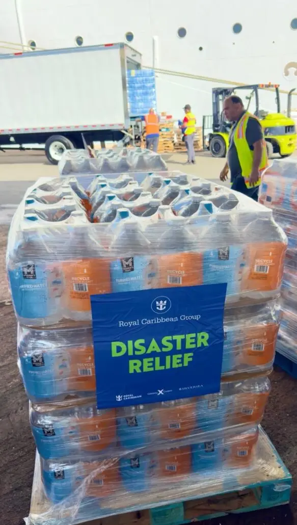 Stacks of bottled water wrapped in plastic are labeled "Royal Caribbean Group Disaster Relief." People in high-visibility vests and a forklift load supplies into a truck, supporting the Jamaica relief effort.